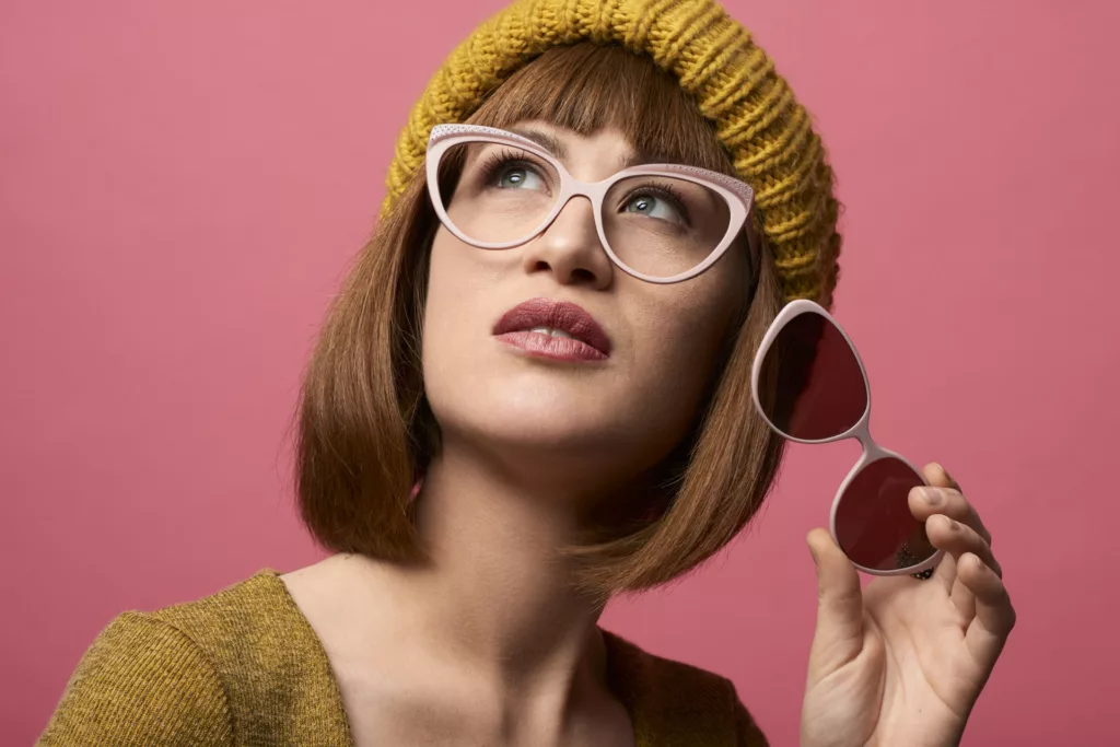 ritratto di giovane donna su sfondo rosa, con cappello color senape, mentre indossa occhiali da vista ed in mano tiene mascherina applicabile sole. Foto realizzata dal fotografo commerciale duzimage a La Spezia